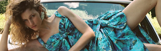 Woman reclining on a car wearing a turquoise floral off-shoulder dress with voluminous sleeves in a fashion editorial setting